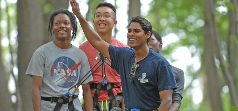 The image shows a group of young people outdoors, likely at an adventure park or similar setting. They are wearing harnesses and appear to be engaged in an activity that involves ropes or climbing. The individuals are smiling and seem to be enjoying themselves. The background is blurred, suggesting a natural environment with trees.